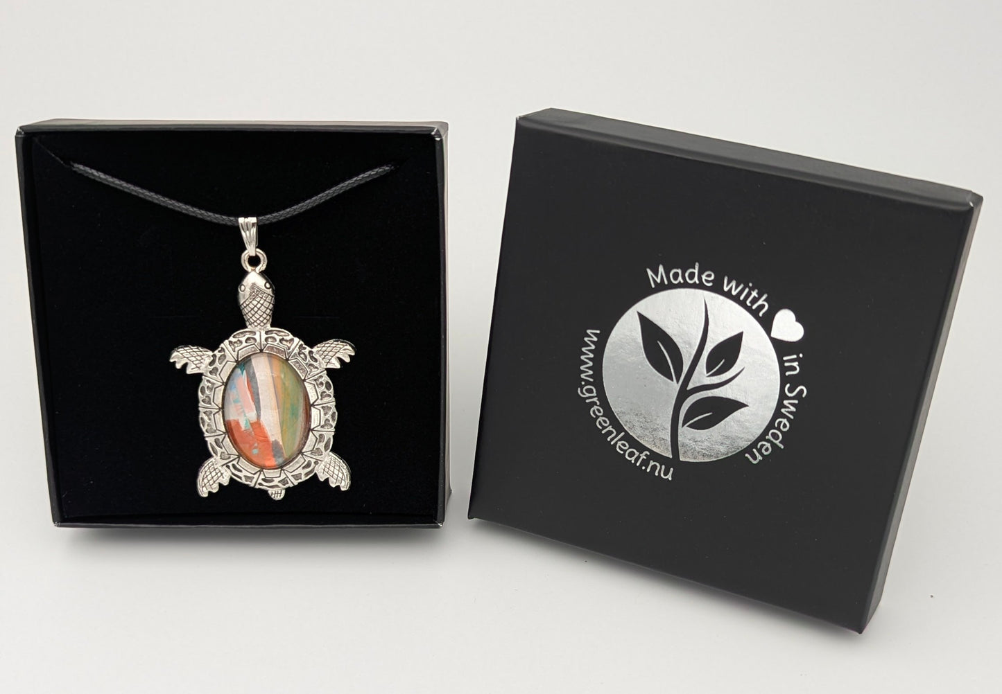 Tortoise-shaped pendant with multicolored stone in a black box, featuring a brand logo.
